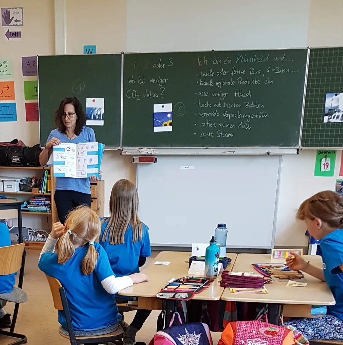Dozentin zeigt der Klasse den Klimaschutz-Vertrag, an der Tafel steht „1, 2 oder 3 – Wo ist weniger CO₂ dabei?“