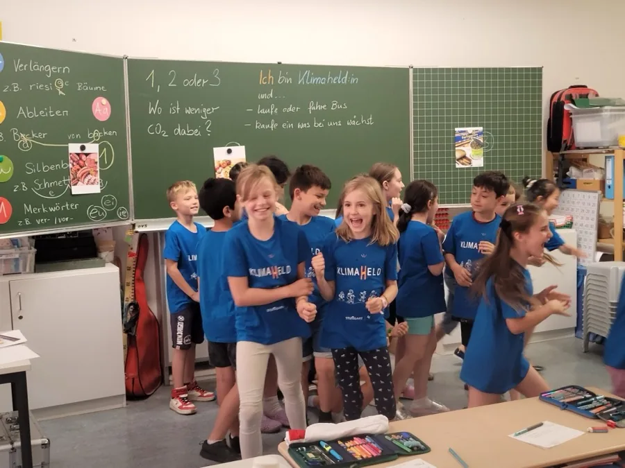 Lachende Kinder in Klimahelden-T-Shirts springen im Klassenzimmer zwischen den Antwortfeldern, an der Tafel steht die Spielfrage